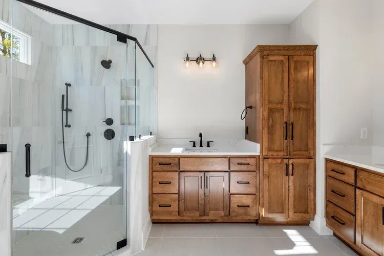 Modern bathroom with glass walk-in shower, wood vanity, and tall cabinet in bright natural light