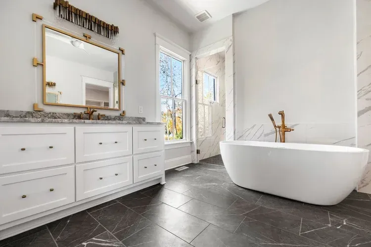 Bright modern bathroom with white vanity, large mirror, and freestanding tub on dark tile floor