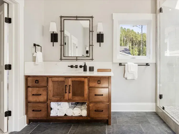Bright bathroom with wood vanity, mirror, sconces, white walls, and a walk-in shower.