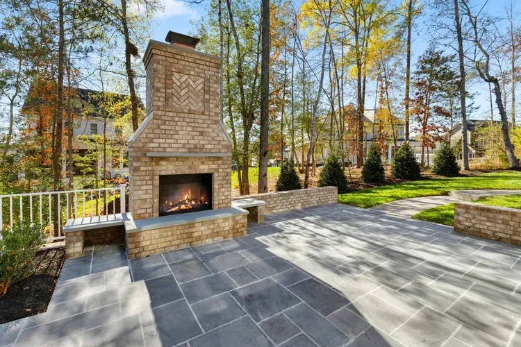 Stone outdoor fireplace on a patio with slate tiles, bordered by autumn trees and a brick retaining wall