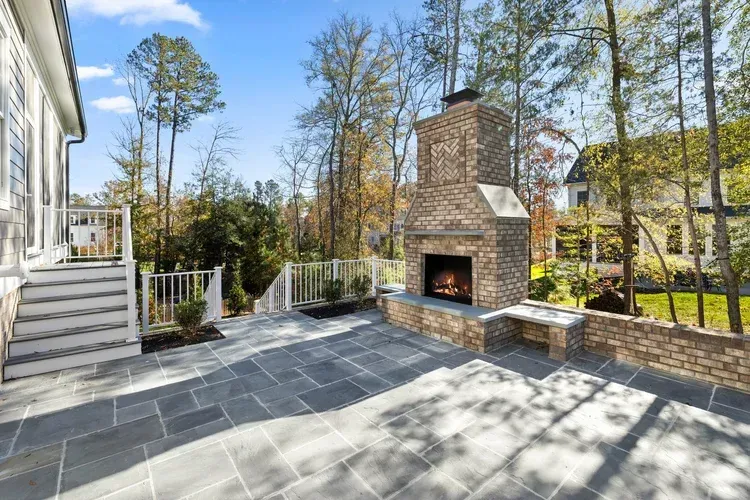 Spacious stone patio with brick fireplace, white railing, and wooded backyard in bright daylight