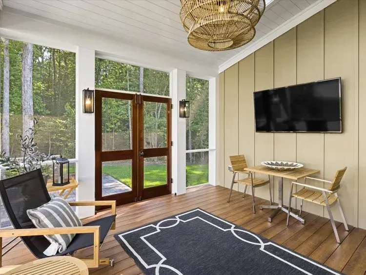 Bright screened porch with wood floor, black rug, wicker chairs, wall TV, and forest view through tall windows