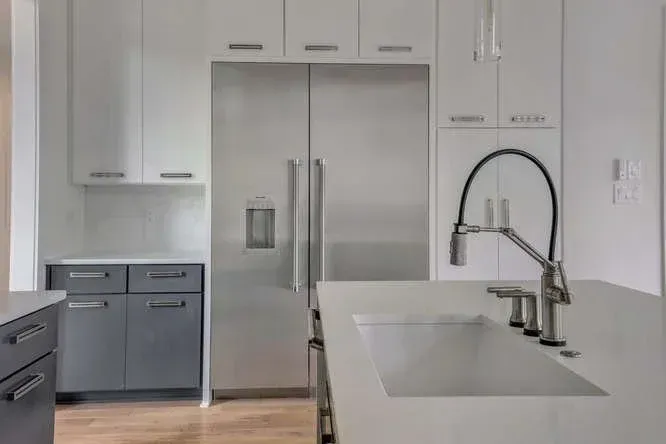 Modern white kitchen with stainless steel refrigerator, gray lower cabinets, and a large island sink.