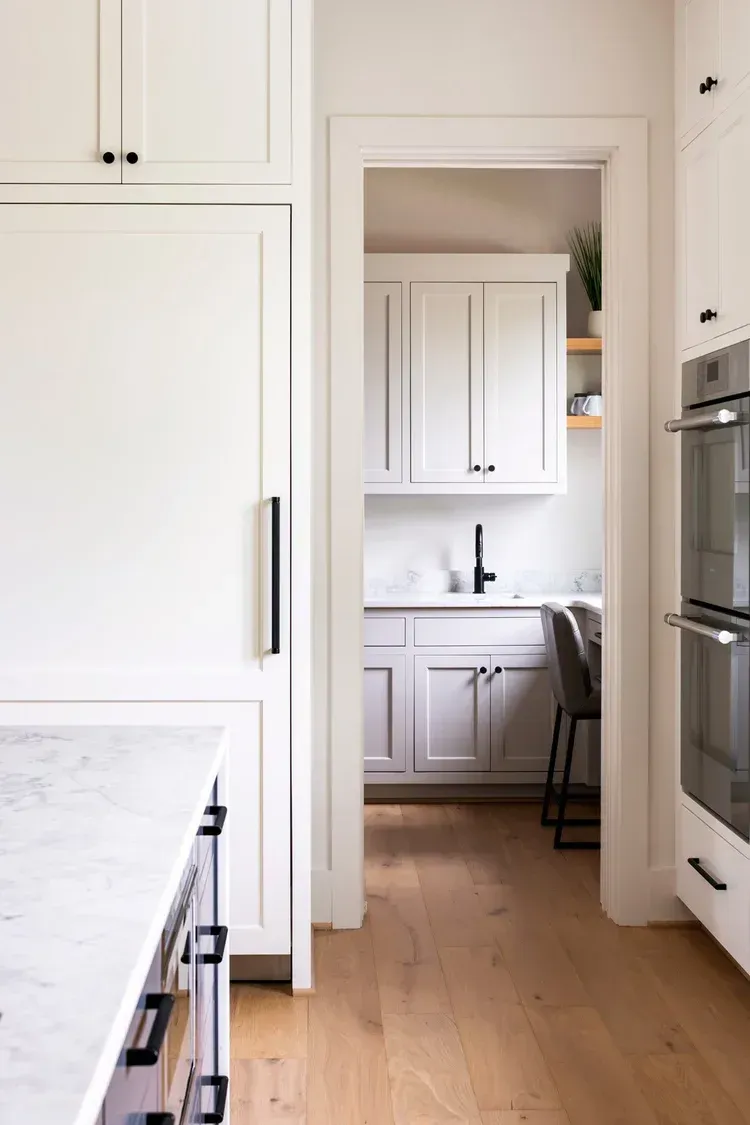 Bright white kitchen with wooden floor, doorway to a small sink area, and built-in ovens on the right