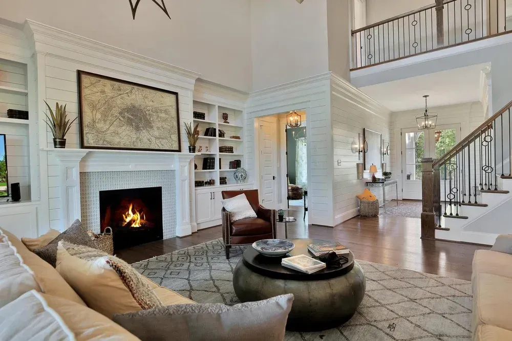 Bright living room with a lit fireplace, beige sofa, round ottoman, and staircase in the background