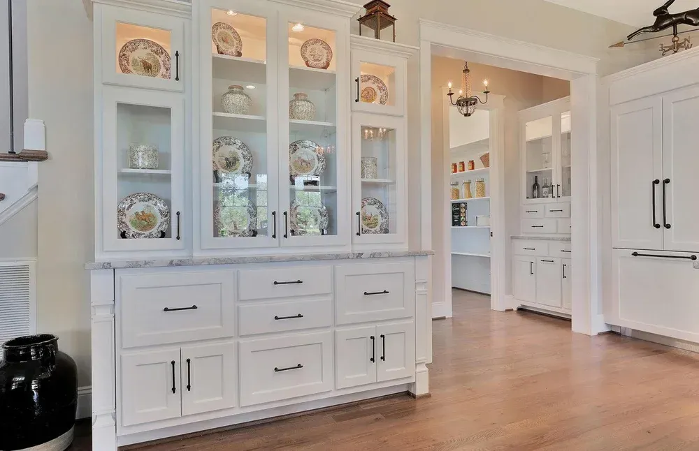 Bright dining room with white china cabinet, hardwood floors, and an open doorway to a kitchen.