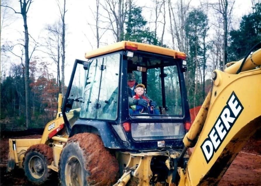 A person and a child sit inside the cab of a yellow John Deere backhoe in a wooded area.