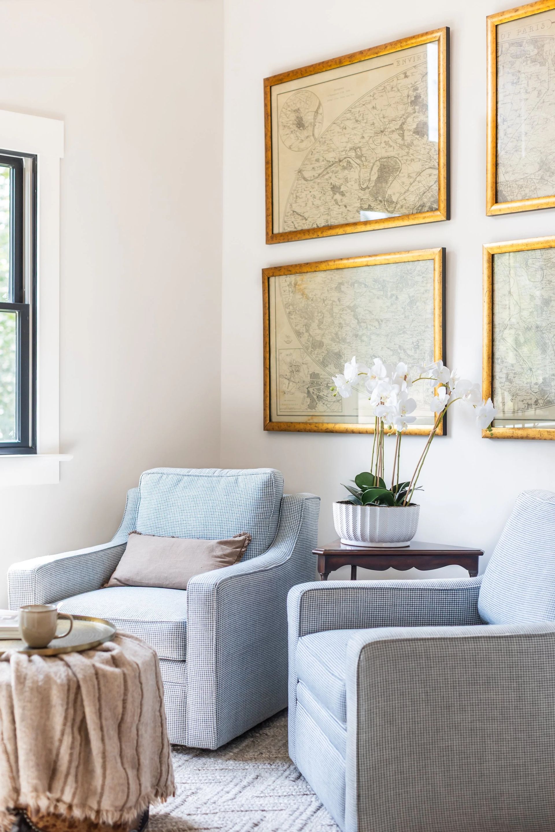 Two light blue armchairs sit in a bright room, featuring four framed antique maps on the wall and a small potted orchid.