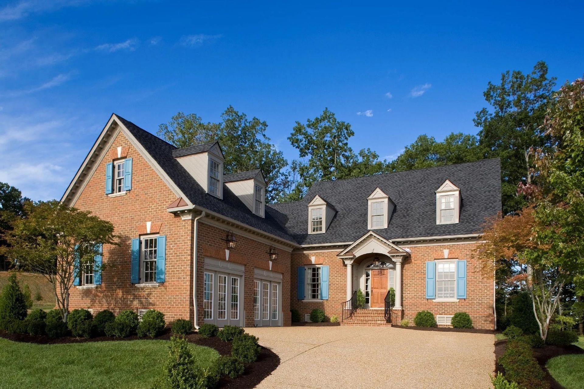 A two-story brick home with a gravel driveway, blue shutters, and a dormer roofline under a bright blue sky.