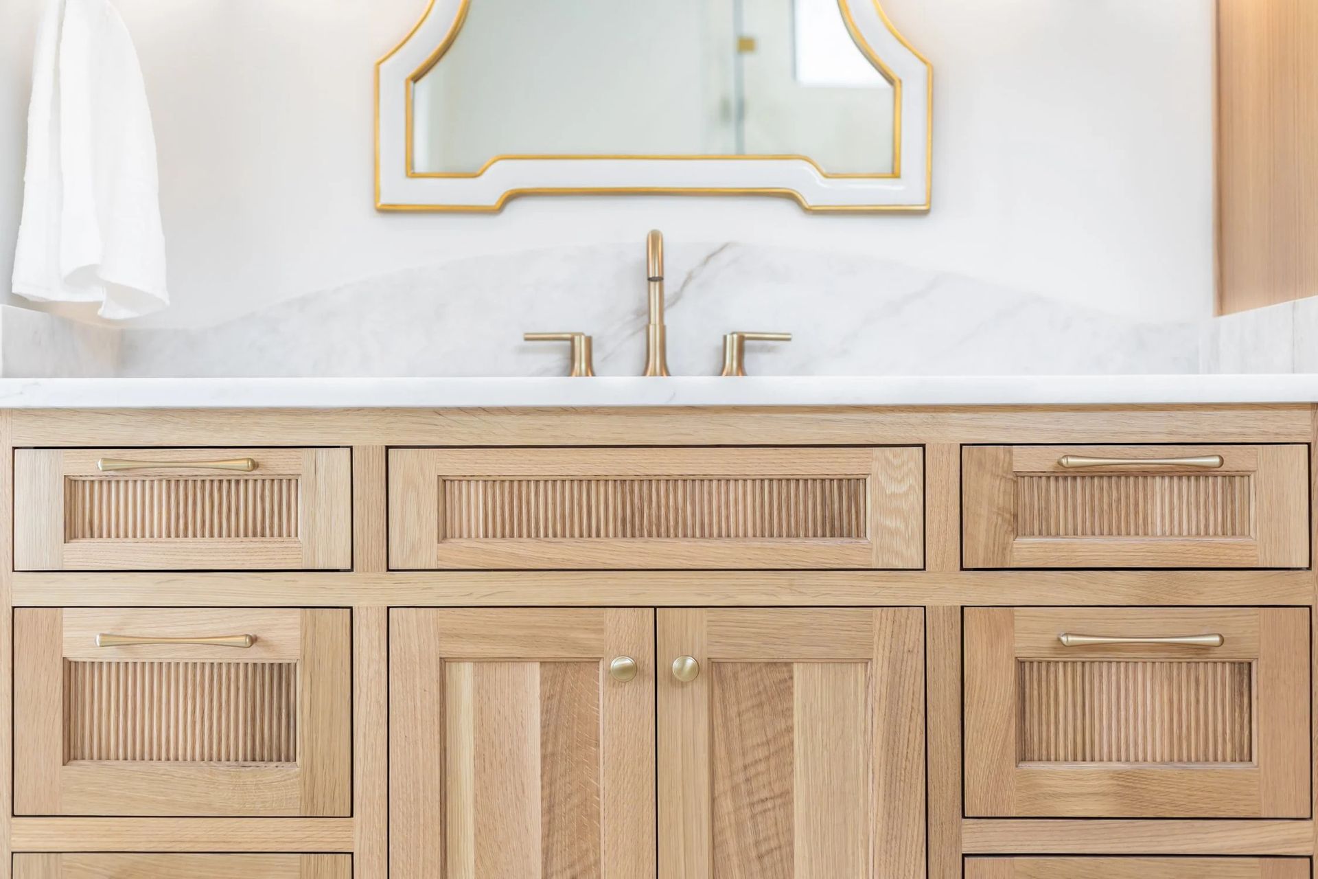 A bathroom vanity with light wood cabinetry, gold fixtures, a white marble countertop, and a uniquely shaped mirror.