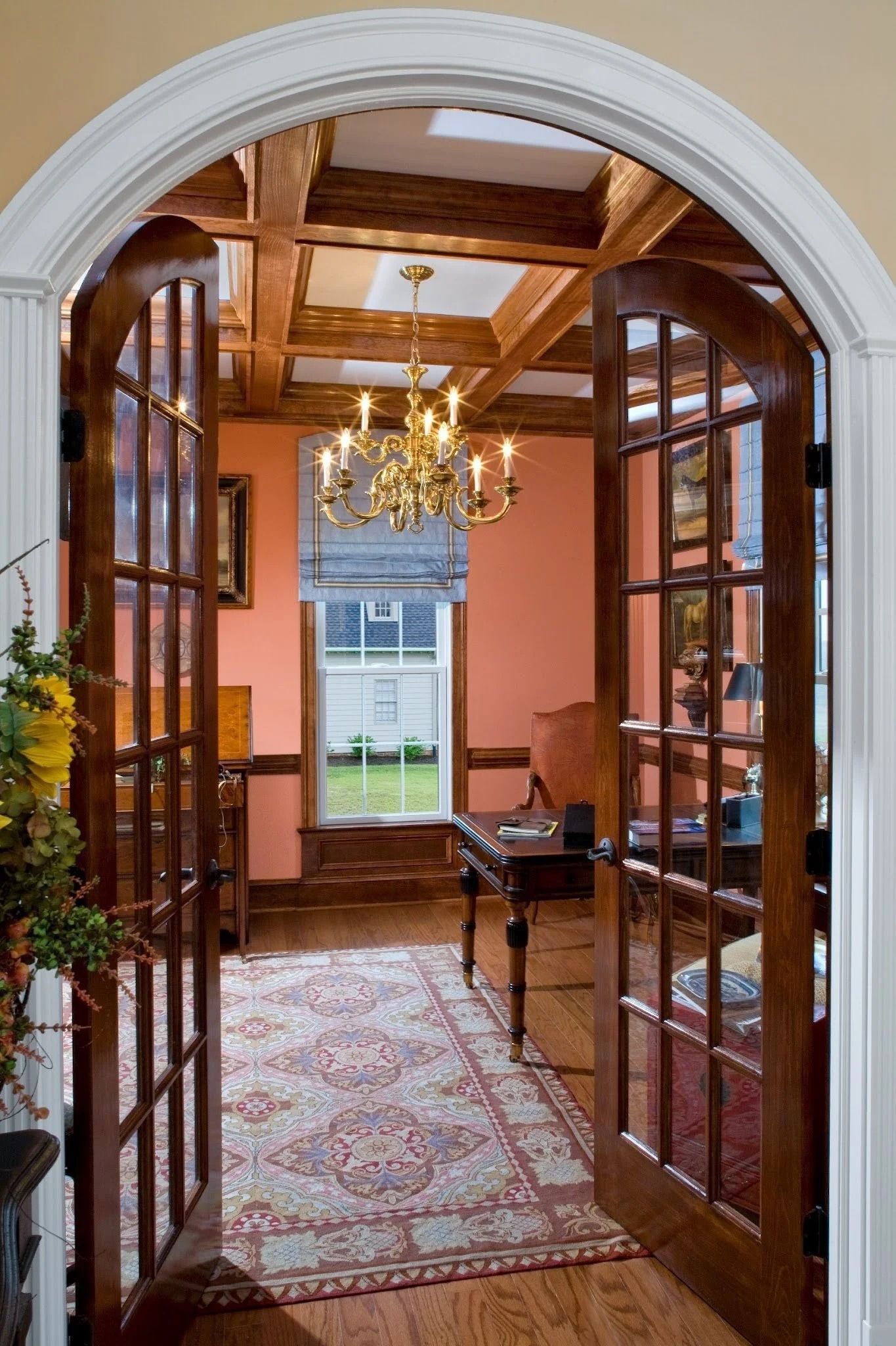 French doors open into a home office with coral walls, a coffered ceiling, a brass chandelier, and an area rug.