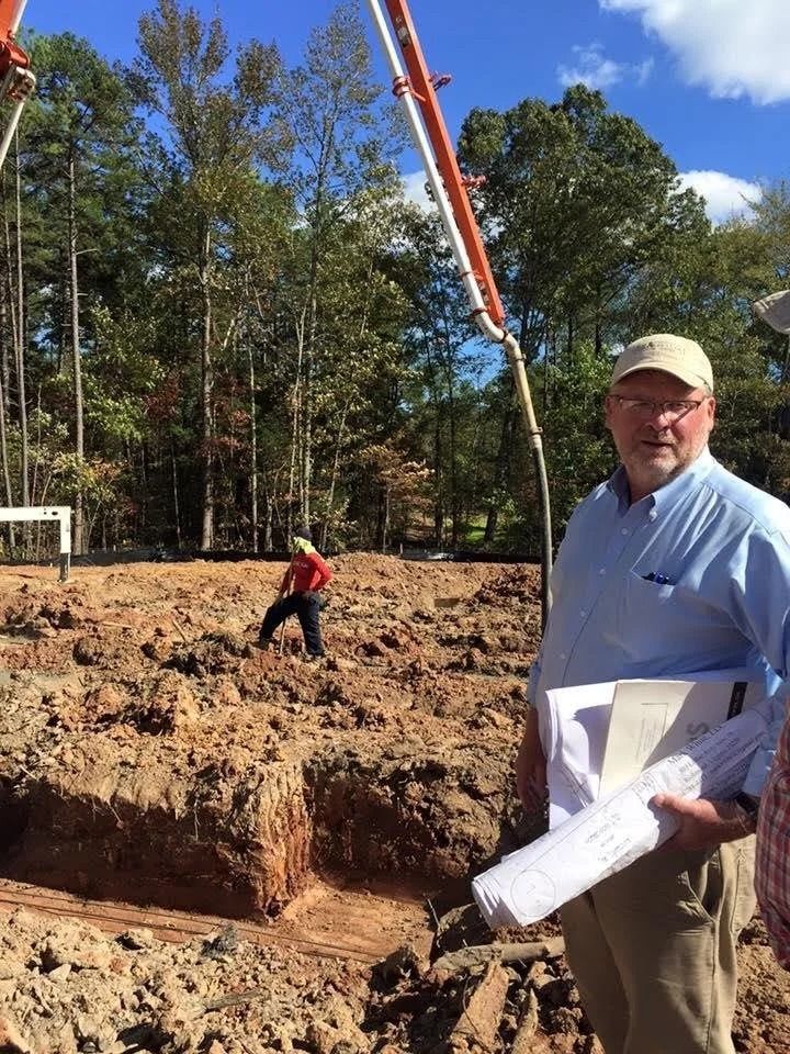 A man holding construction plans stands at a dirt excavation site with a concrete pump in the background.