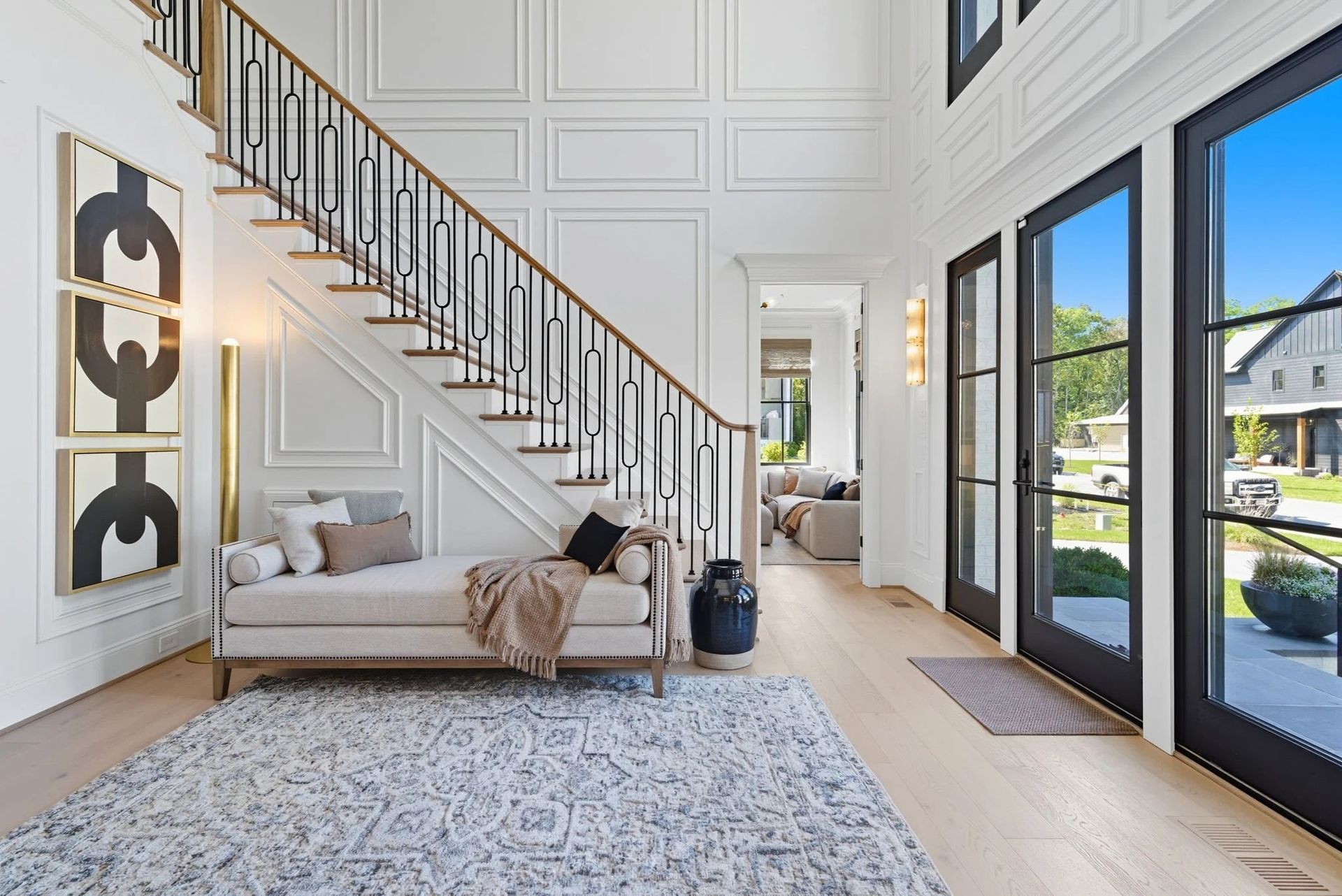 A modern foyer with a white staircase, a daybed under the stairs, a textured rug, and tall black-framed glass doors.