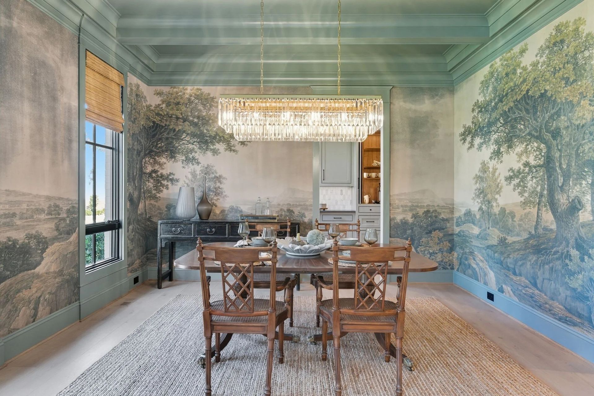 A formal dining room features a mural of a landscape on the walls, a wooden table with chairs, and a crystal chandelier.