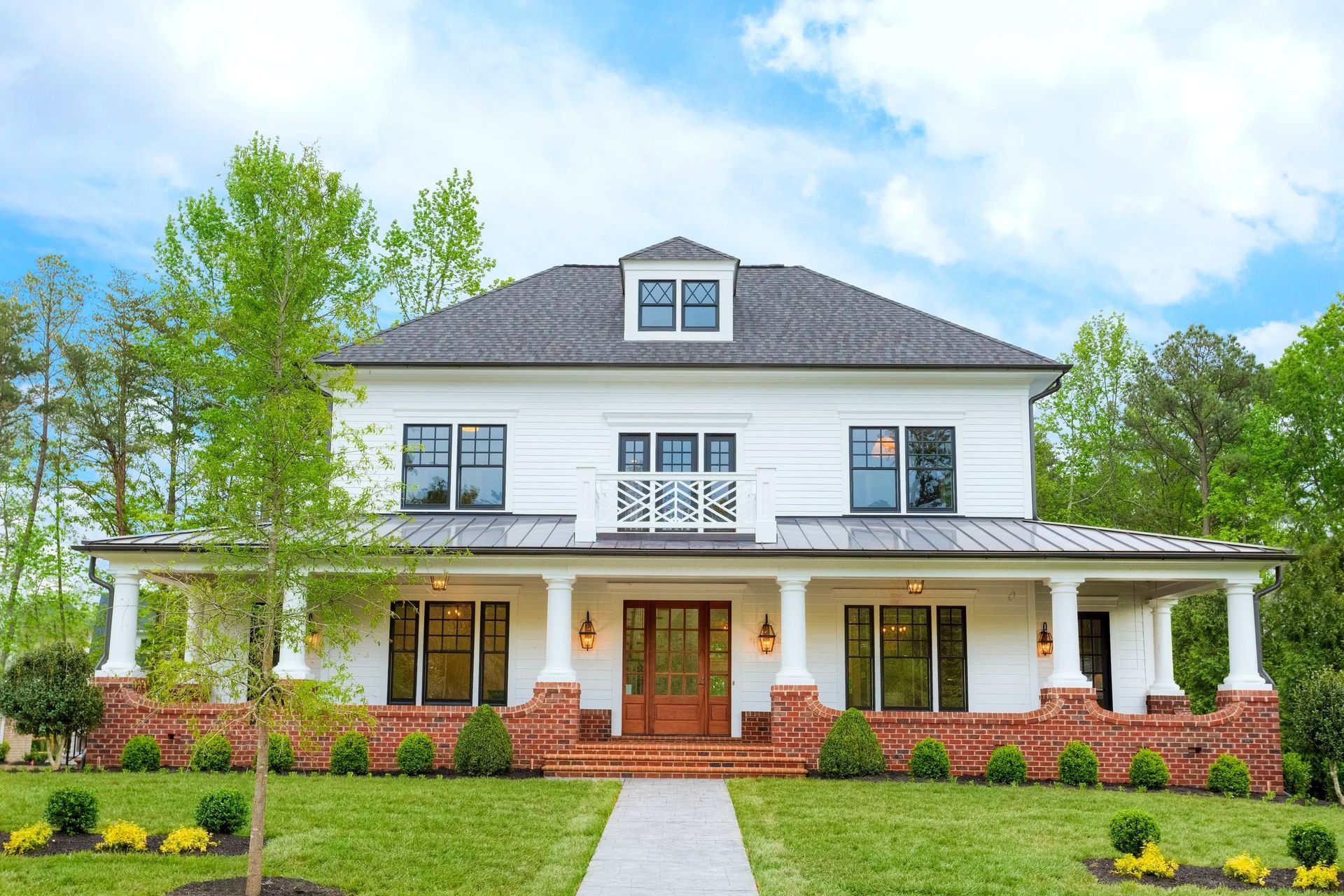 A two-story white house with a dark roof, a brick porch base, wrap-around porch, and surrounding trees under a blue sky.