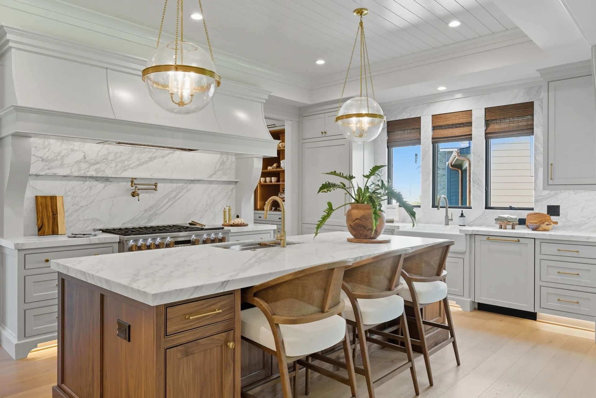 A modern white kitchen featuring a large wood island with seating, marble countertops, pendant lights, and a hood vent.