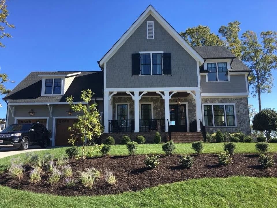 A two-story suburban home with a gray exterior, stone lower level, gabled roof, covered porch, and a black car parked.