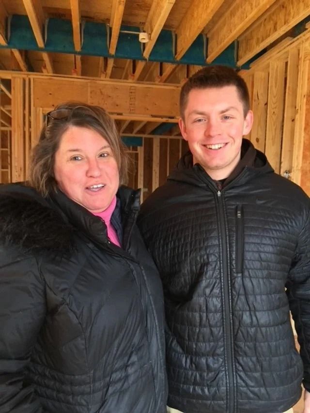 A smiling woman and man in winter jackets stand together inside a house under construction with visible wooden beams.