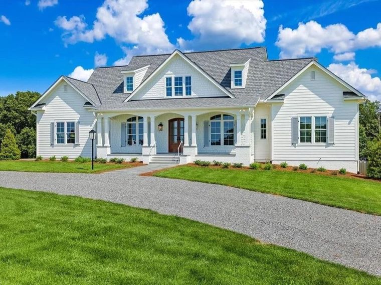 A white, farmhouse-style house with a gray roof and a gravel driveway sits on a green lawn under a blue, cloudy sky.