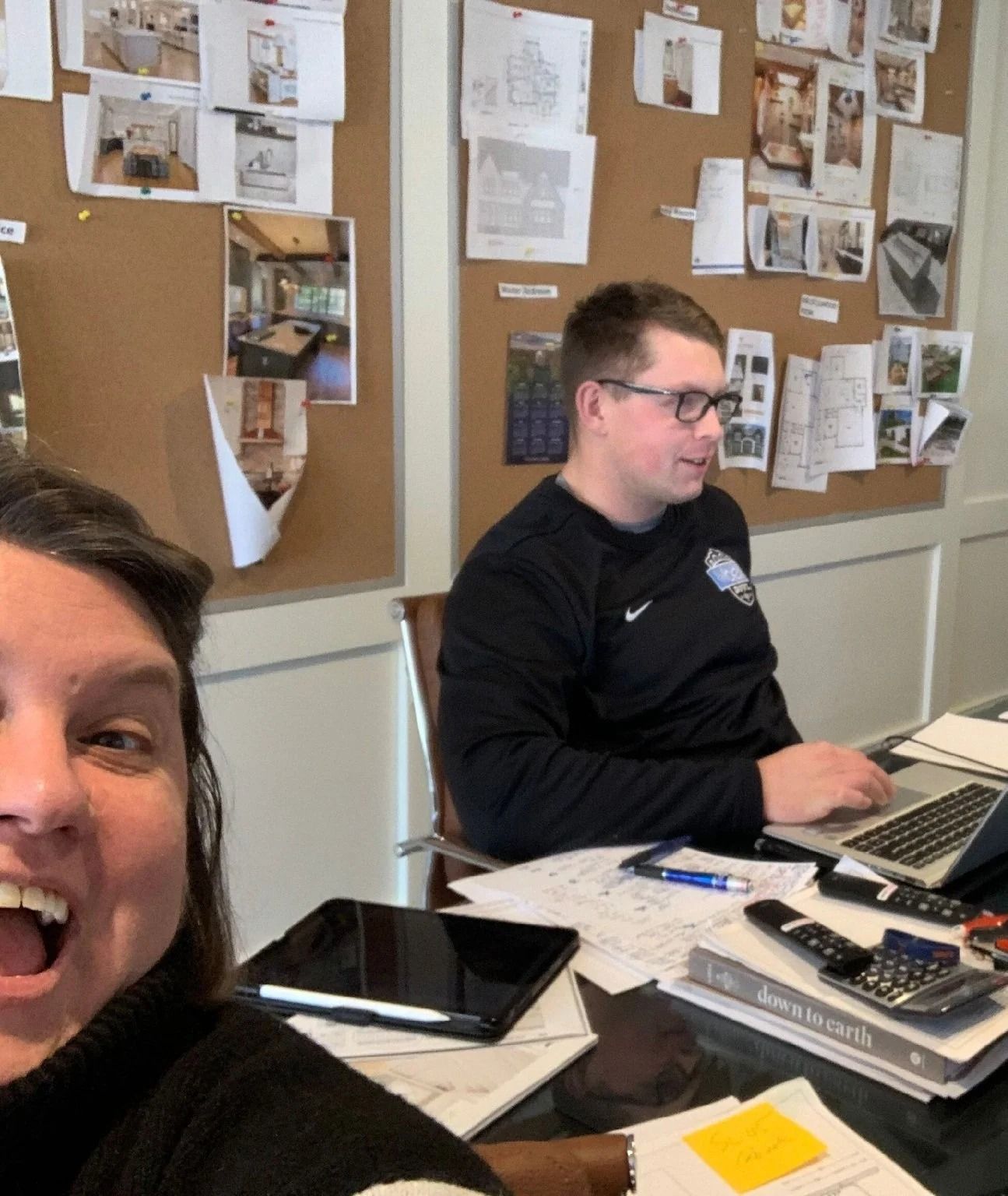 A person smiles for a selfie while a colleague works on a laptop at a desk in front of a bulletin board full of papers.