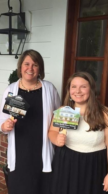 Two people stand side-by-side outdoors, smiling and holding house-shaped promotional fans featuring photos of homes.