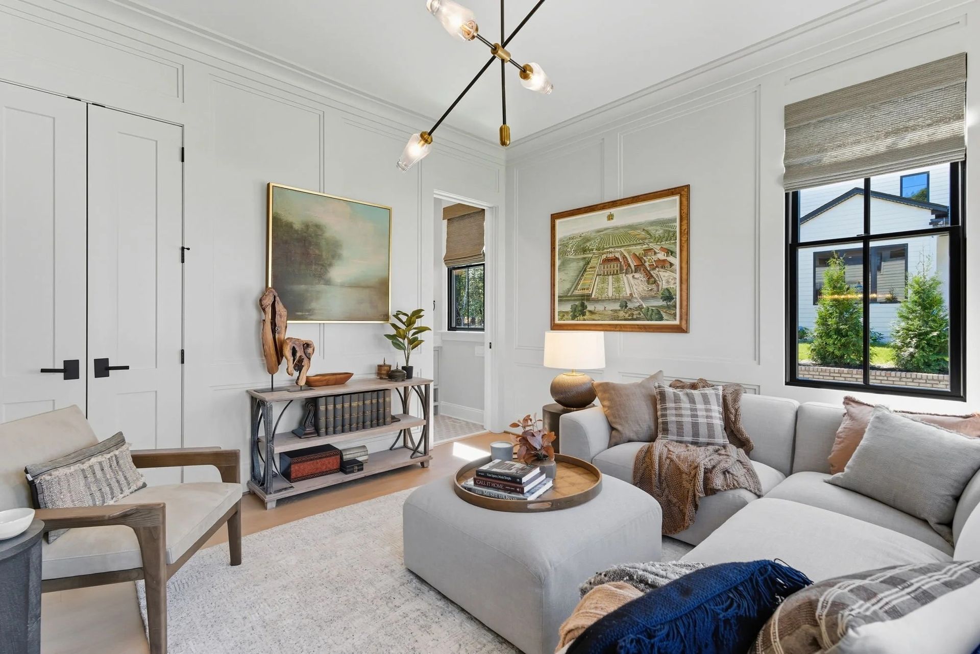 A bright, modern living room with white paneled walls, a gray sectional sofa, a large ottoman, and an armchair.