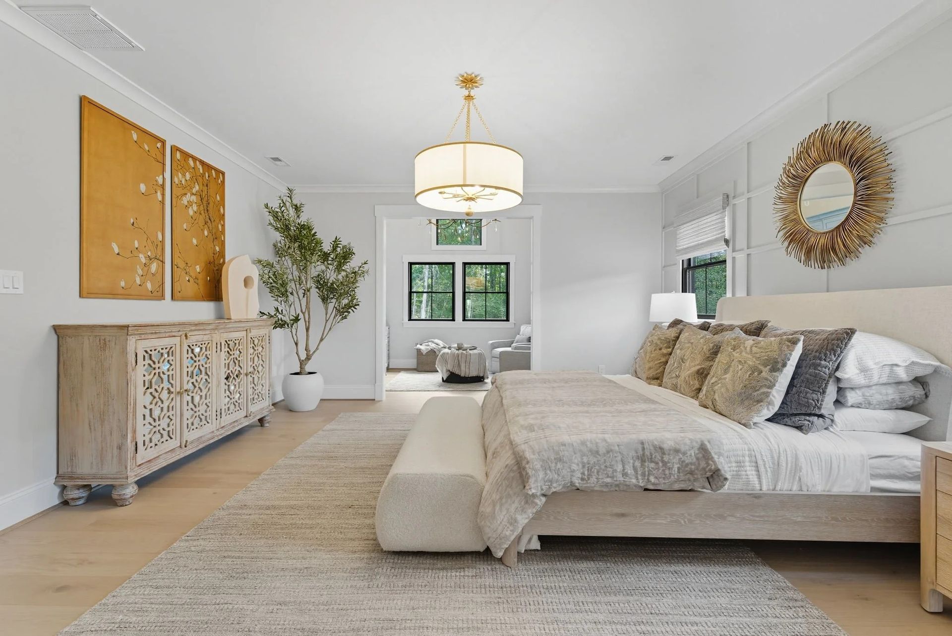 A bright, airy bedroom featuring a wooden bed, patterned bedding, a carved sideboard, a potted plant, and a gold mirror.