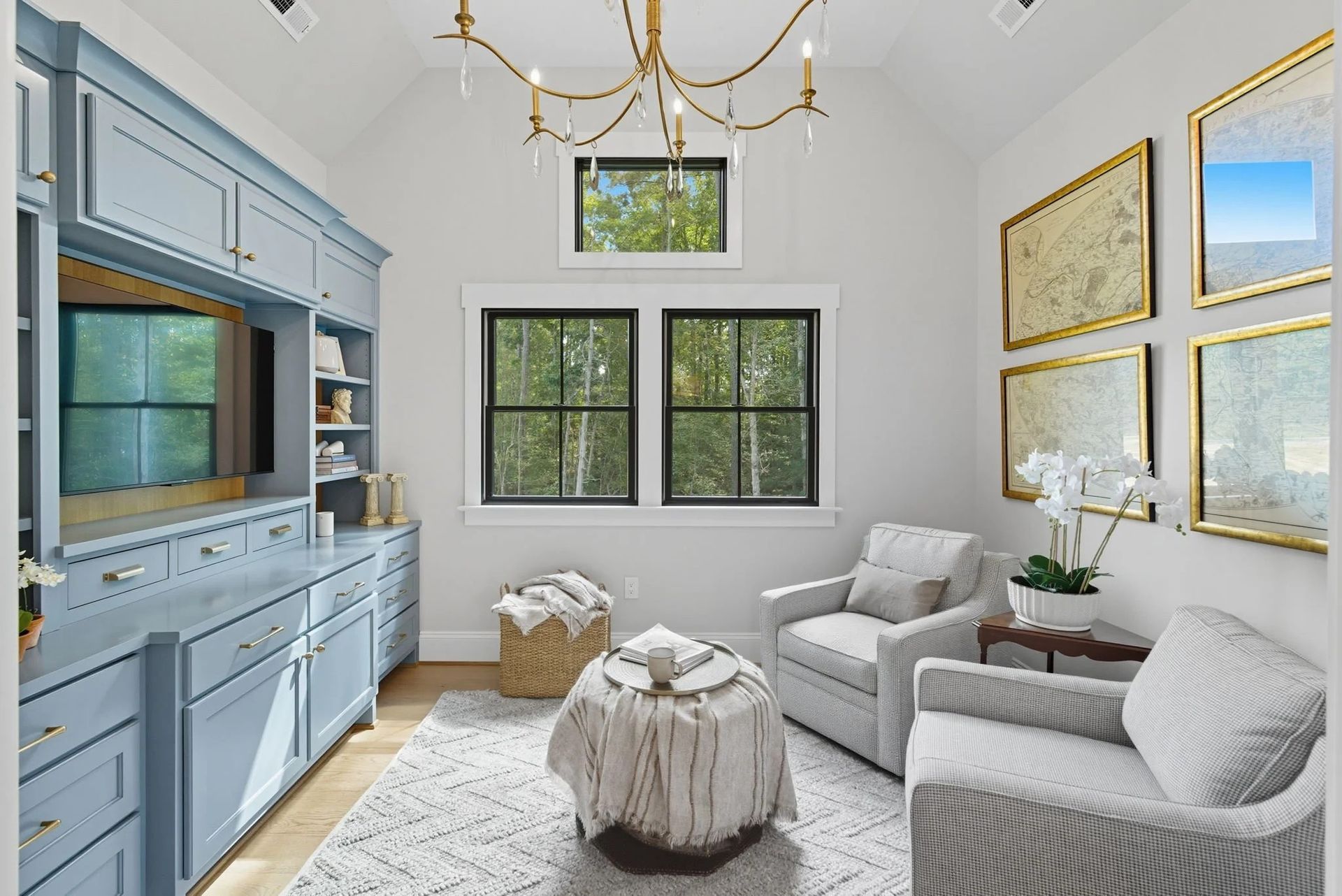 A cozy, light-filled sitting room featuring blue cabinetry, two gray armchairs, a central ottoman, and framed wall art.
