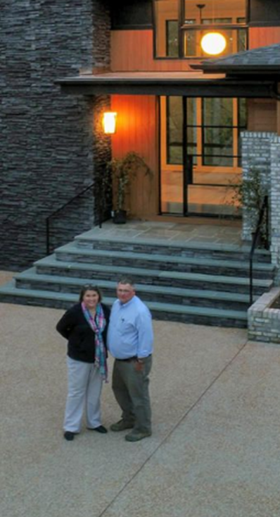 A couple stands on a paved driveway in front of the stone-walled entrance of a modern home with glowing lights.