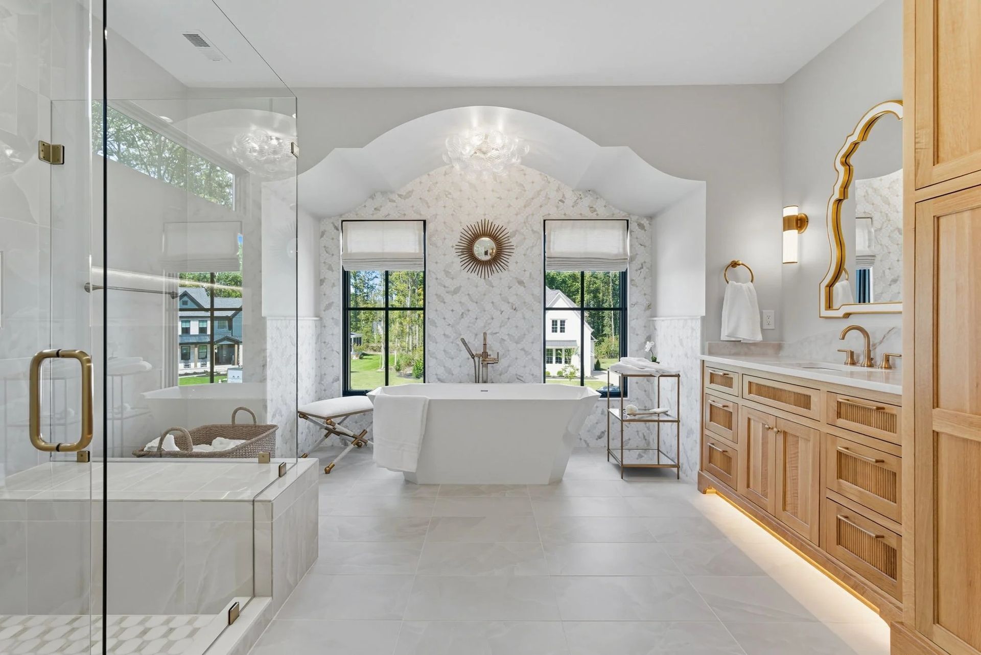 A modern bathroom with a freestanding tub, glass shower, natural wood vanity, and arched alcove with tiled walls.