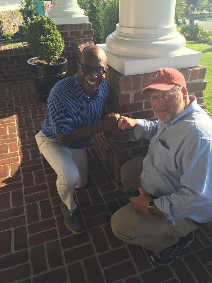 Two people kneel on a brick patio by a white column, smiling and sharing a fist bump.