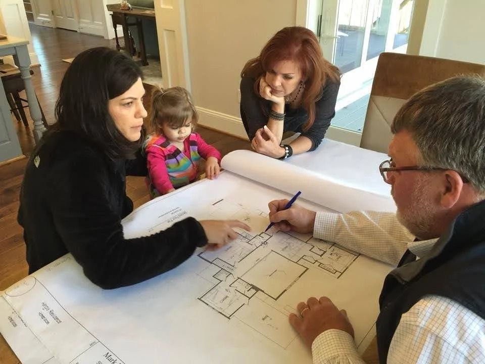 Two women and a small child observe a person using a blue pen to mark architectural floor plans on a large table.