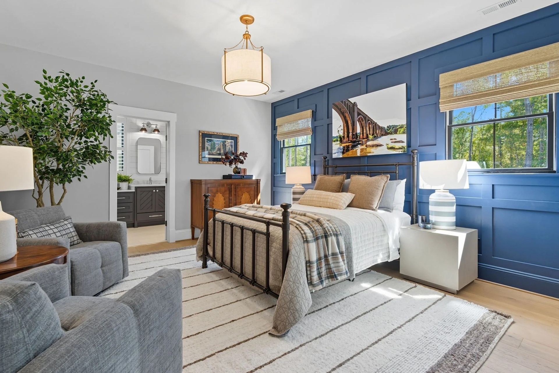 A bedroom with an iron bed, blue wainscoting, two gray armchairs, and light wood floors.