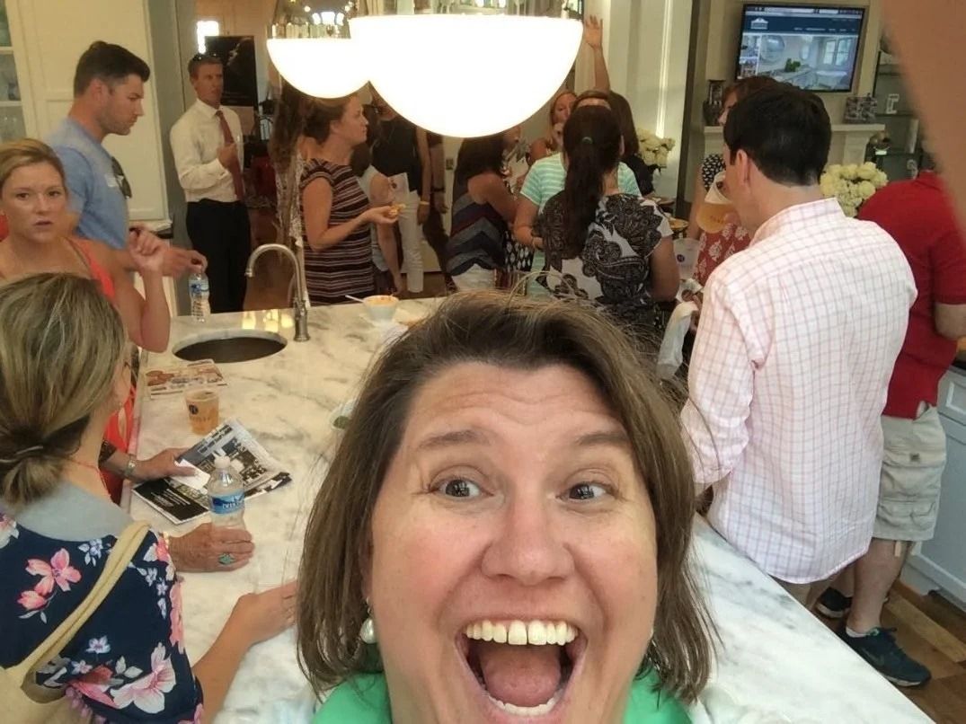 A woman smiles broadly for a selfie at a social gathering in a kitchen with several people mingling in the background.