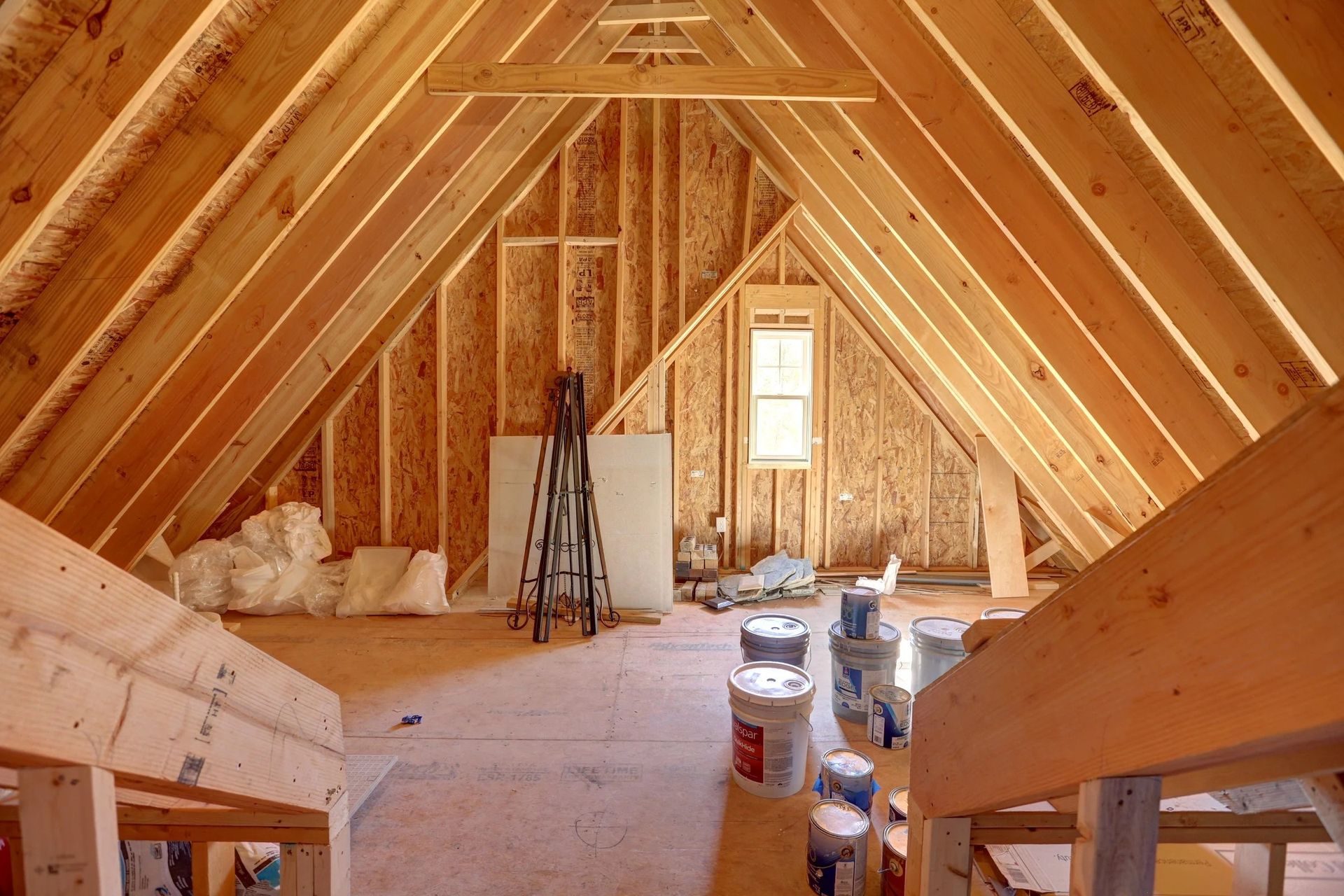 An unfinished attic space under construction with exposed wooden rafters, plywood walls, a small window, and supplies.