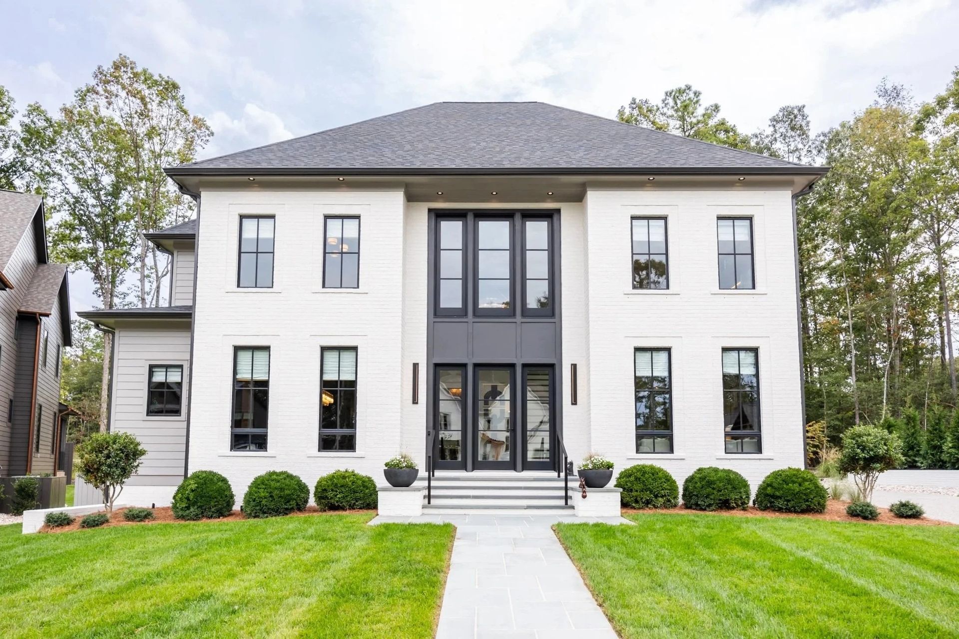 A two-story, white-brick suburban house with a central dark-framed entryway, black windows, and a manicured green lawn.