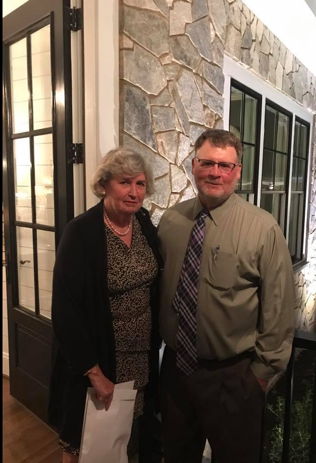 A smiling woman and a man in a dress shirt and tie stand together indoors in front of a stone wall and glass doors.