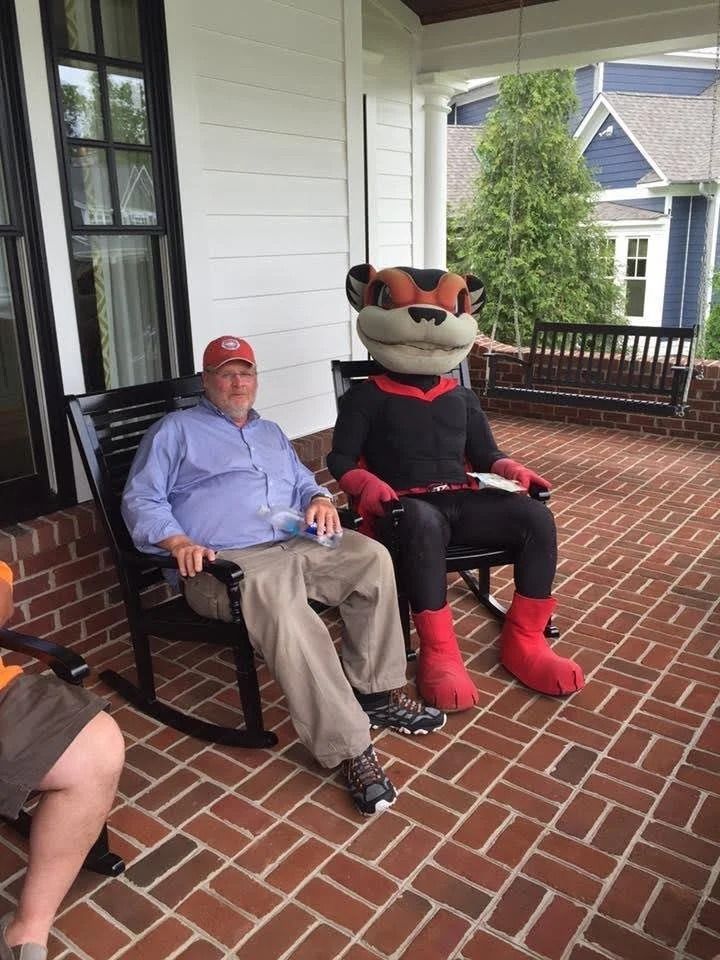An older man and a university mascot sit in rocking chairs on a brick porch.