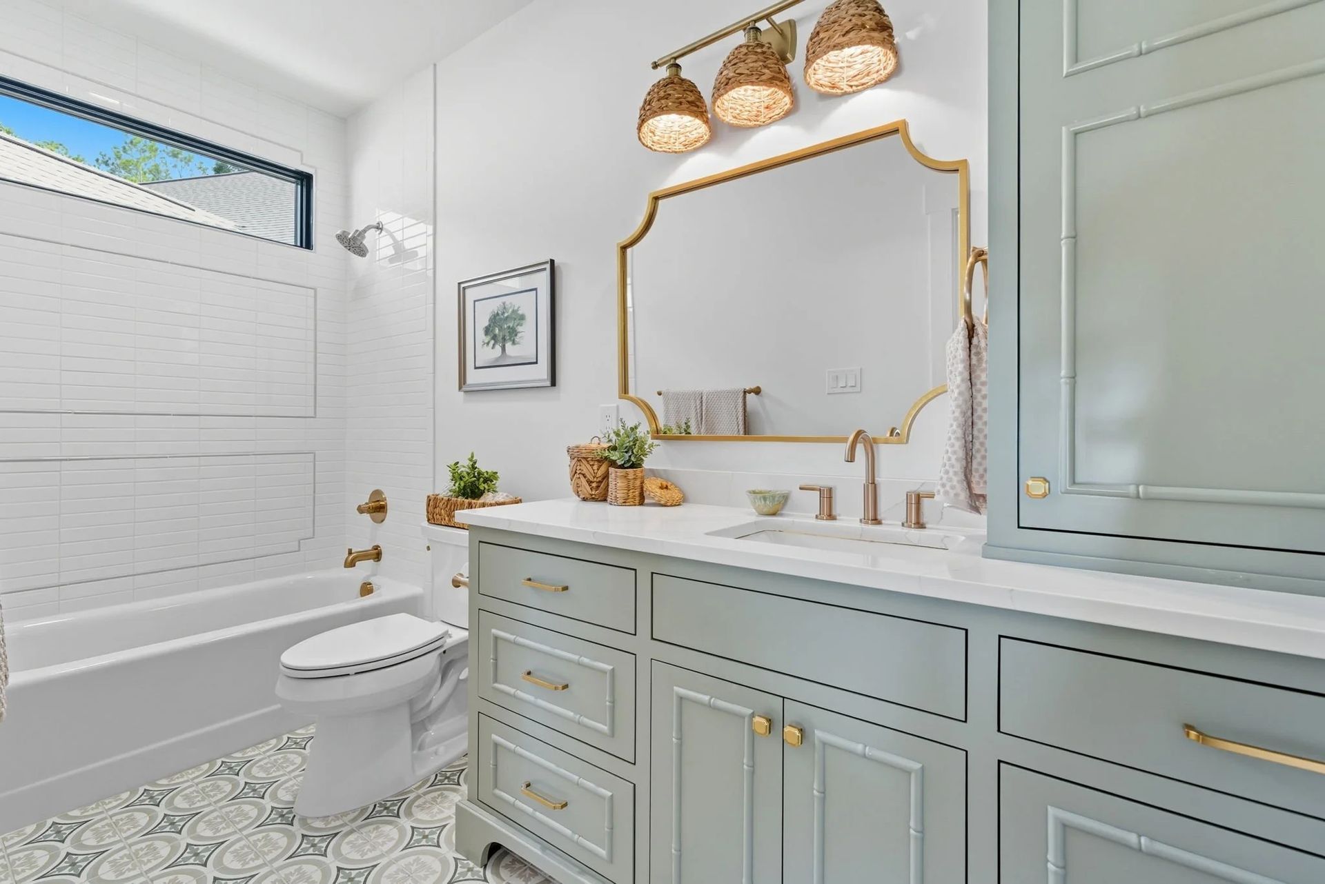 A bright bathroom with sage green cabinets, a white marble countertop, a gold-framed mirror, and patterned floor tiles.