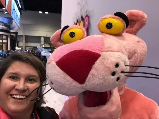 A person smiles while taking a selfie next to a plush Pink Panther mascot character.
