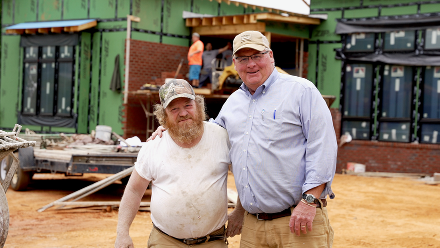 Two men stand smiling with an arm around each other in front of a house under construction with exposed green siding.