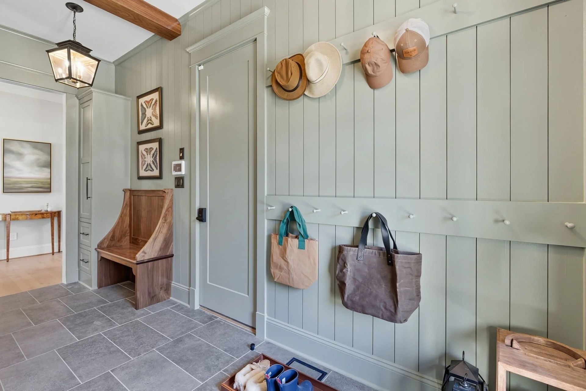 A mudroom featuring sage green vertical paneling, coat hooks with hats and bags, a wood bench, and gray tile flooring.