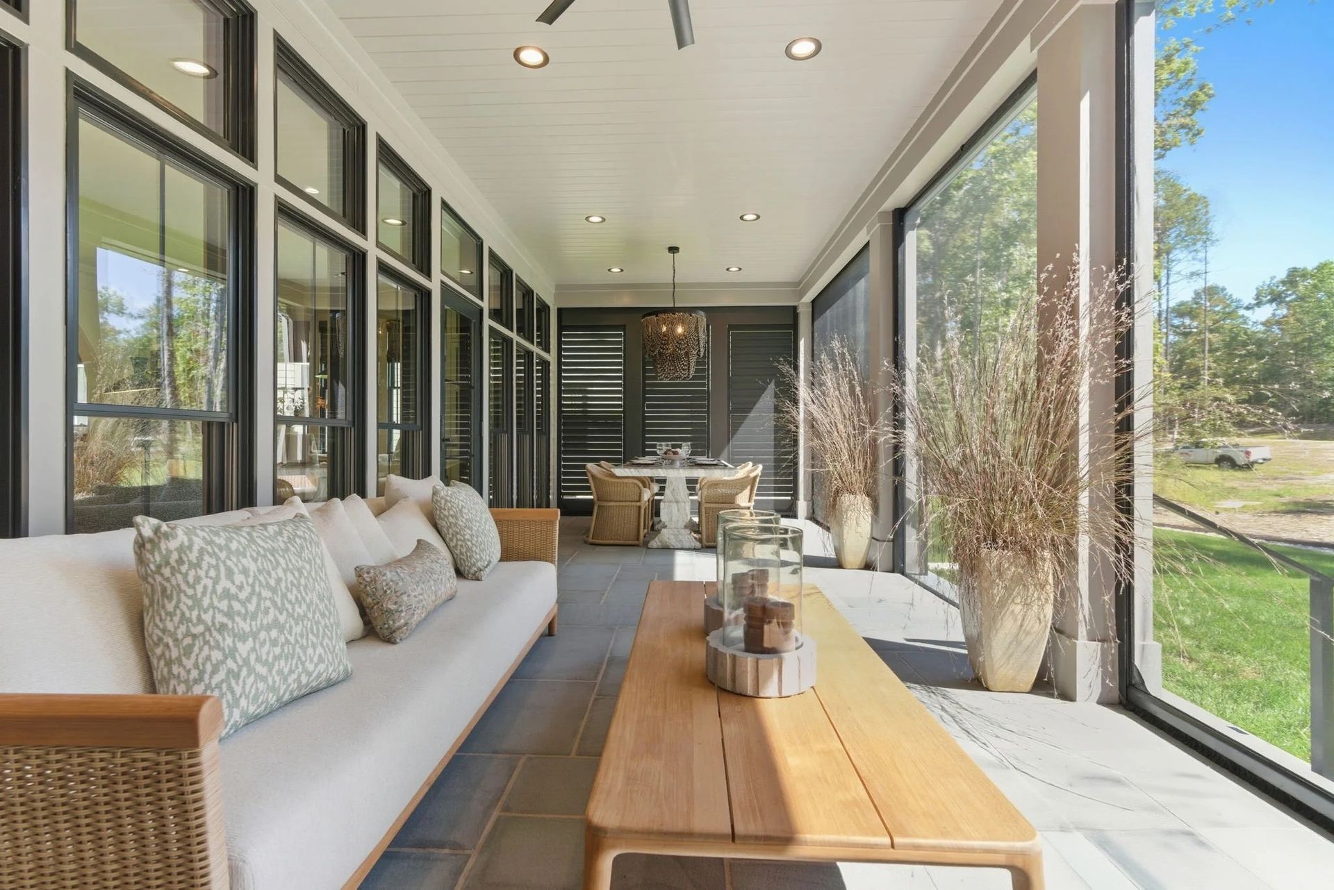 A bright, screened-in porch with a long light-colored sofa, wooden coffee table, and dining set overlooking a yard.