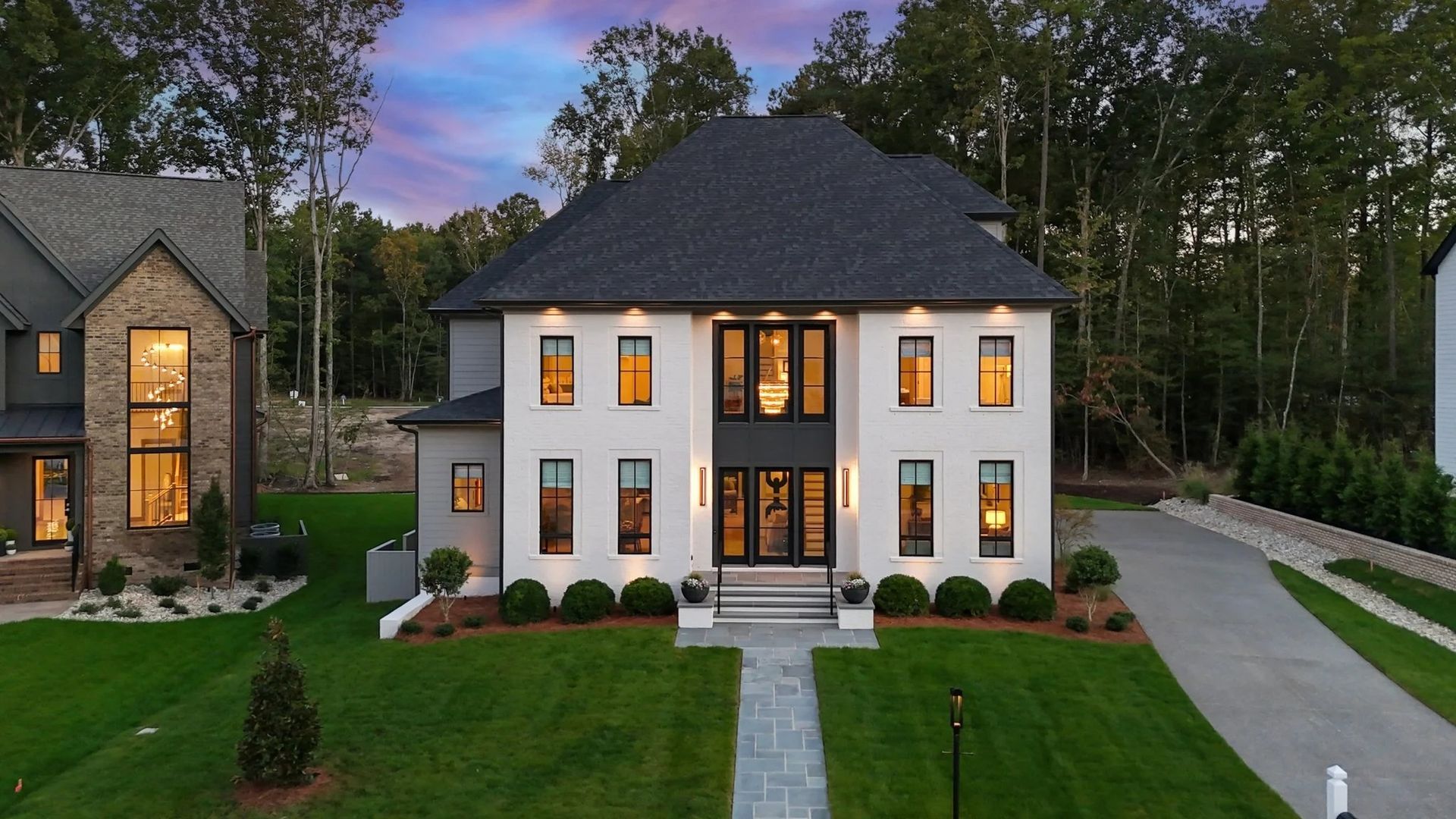 A white brick, two-story house at twilight, with large windows, dark shingled roof, front walkway, and green lawn.
