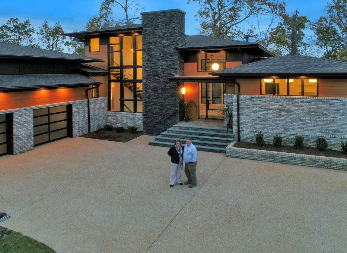 Two people stand on the driveway of a modern home featuring stone accents, large windows, and a multi-level roofline.