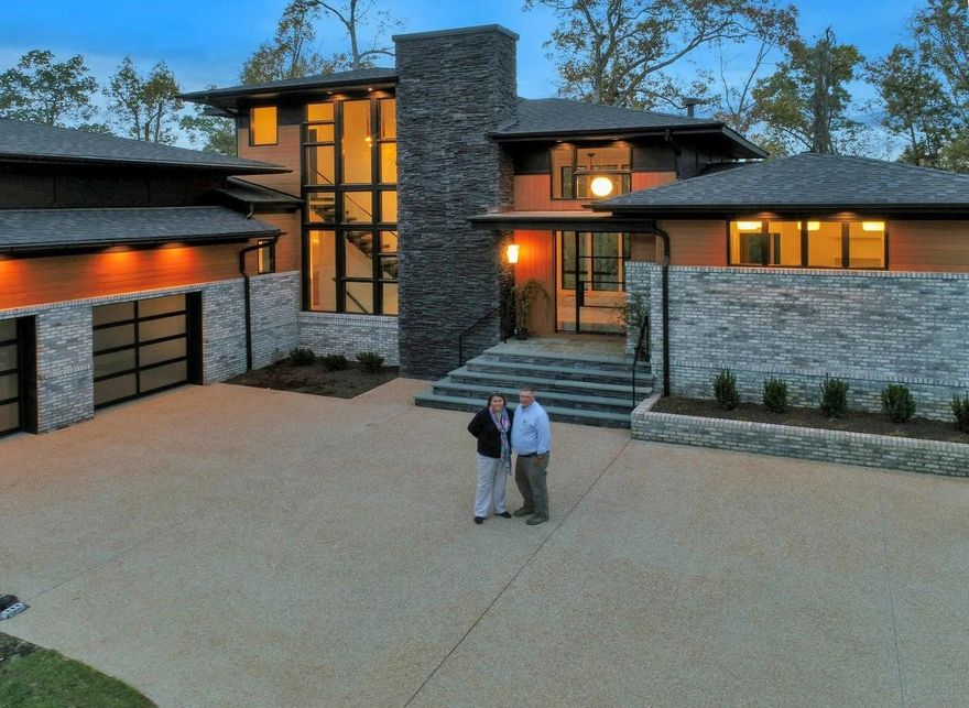 Two people stand in the paved driveway of a modern, multi-level house with stone siding and large glass windows at dusk.