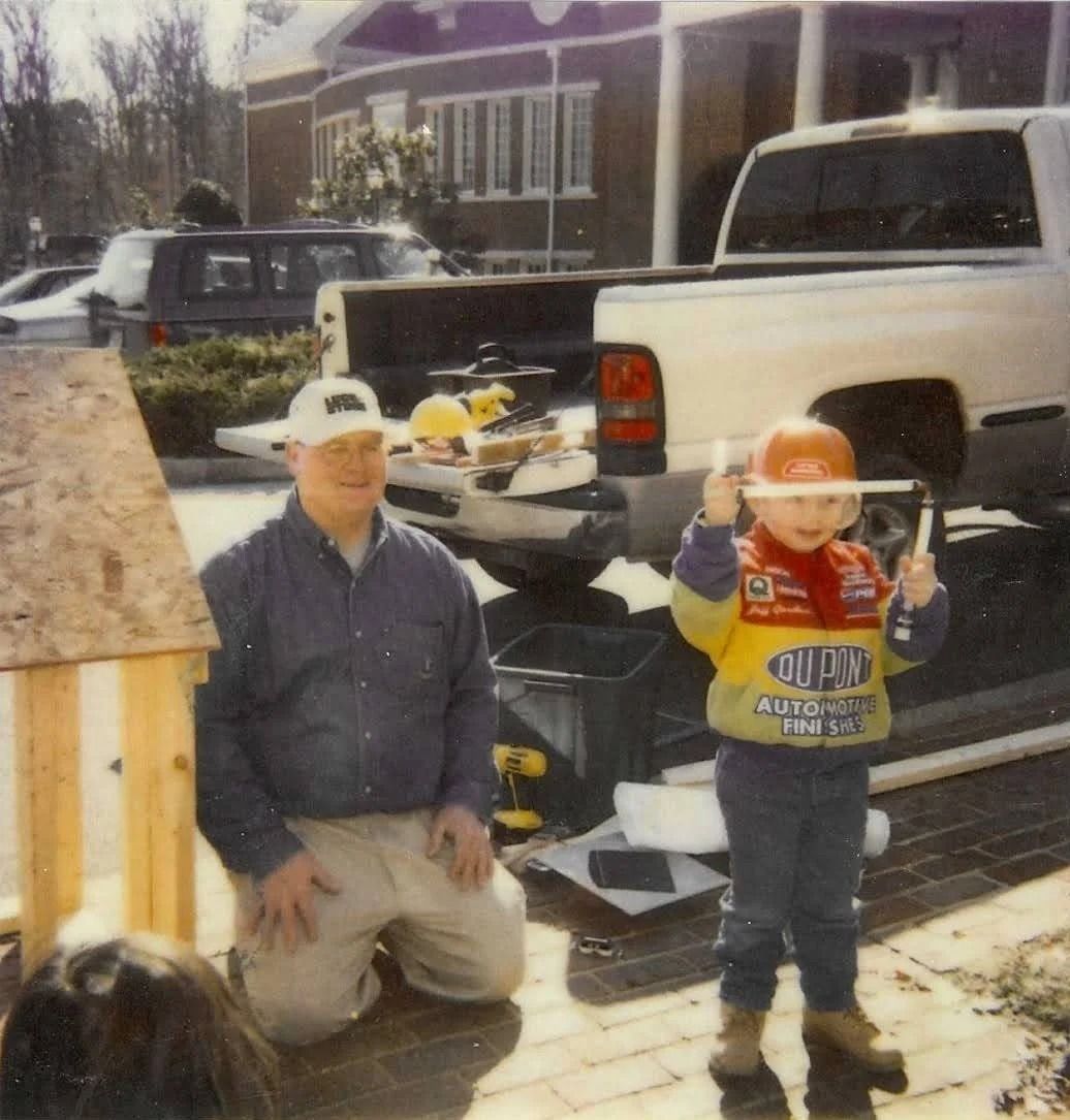 A person kneels next to a child wearing a construction helmet and Dupont jacket in front of a pickup truck and a building.