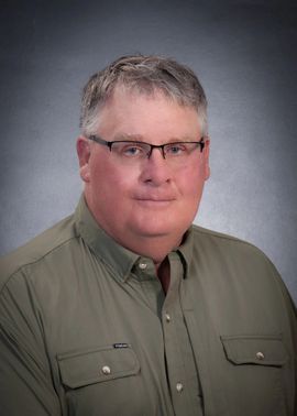 Headshot of a person wearing glasses and a green button-down shirt against a gray background.