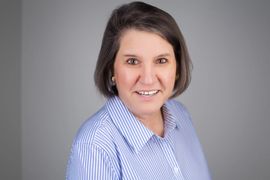 A smiling person with shoulder-length dark hair, wearing a light blue and white pinstriped button-down shirt.