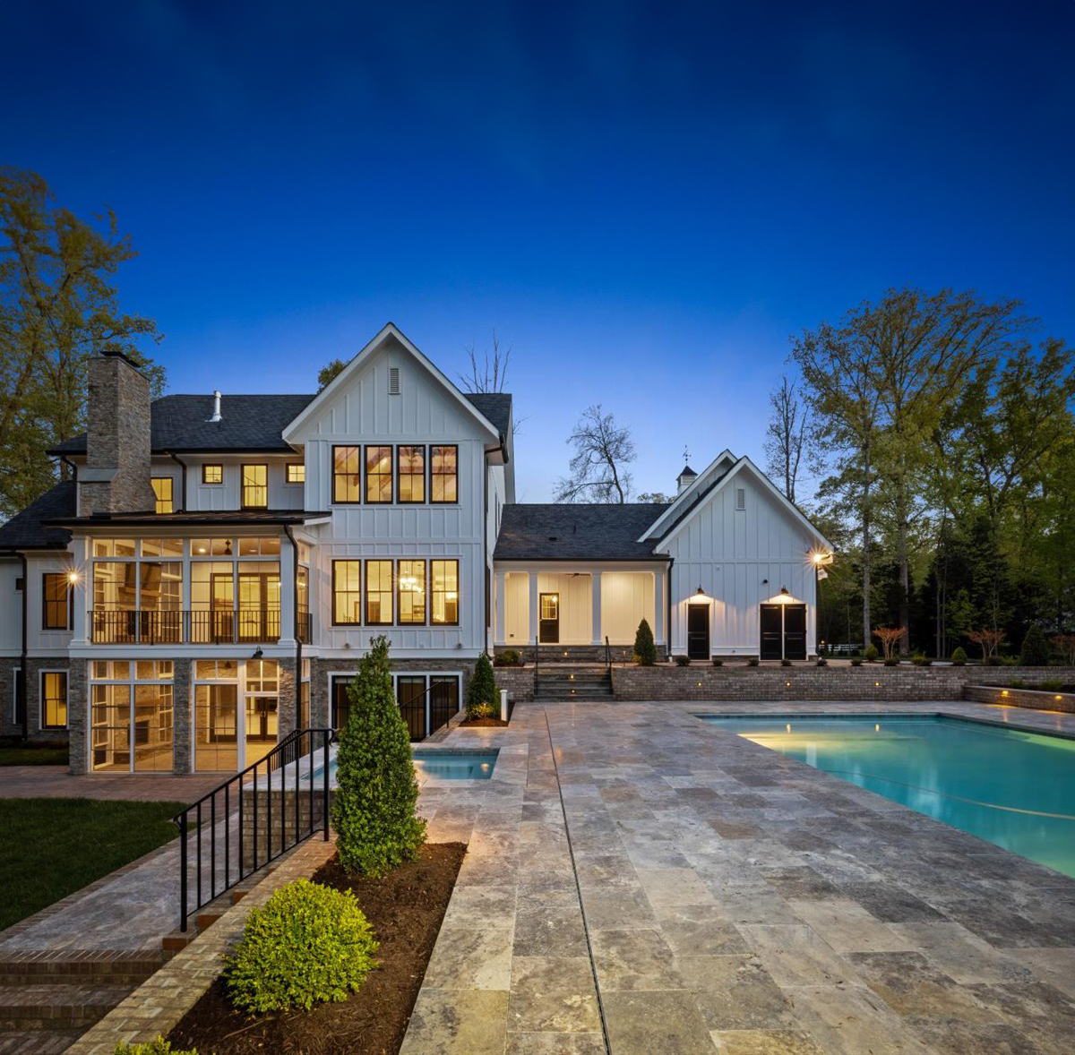 A modern two-story white house with a stone foundation, lit at twilight, overlooking a backyard pool and patio area.